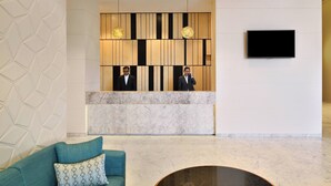 Zona de recepción de diseño moderno, con mostrador de mármol, dos recepcionistas, televisor de pantalla plana y sofá azul turquesa con cojín estampado.