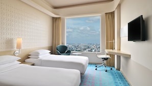 Habitación de hotel con dos camas, un escritorio, una silla, televisión y un ventanal con vistas a la ciudad.
