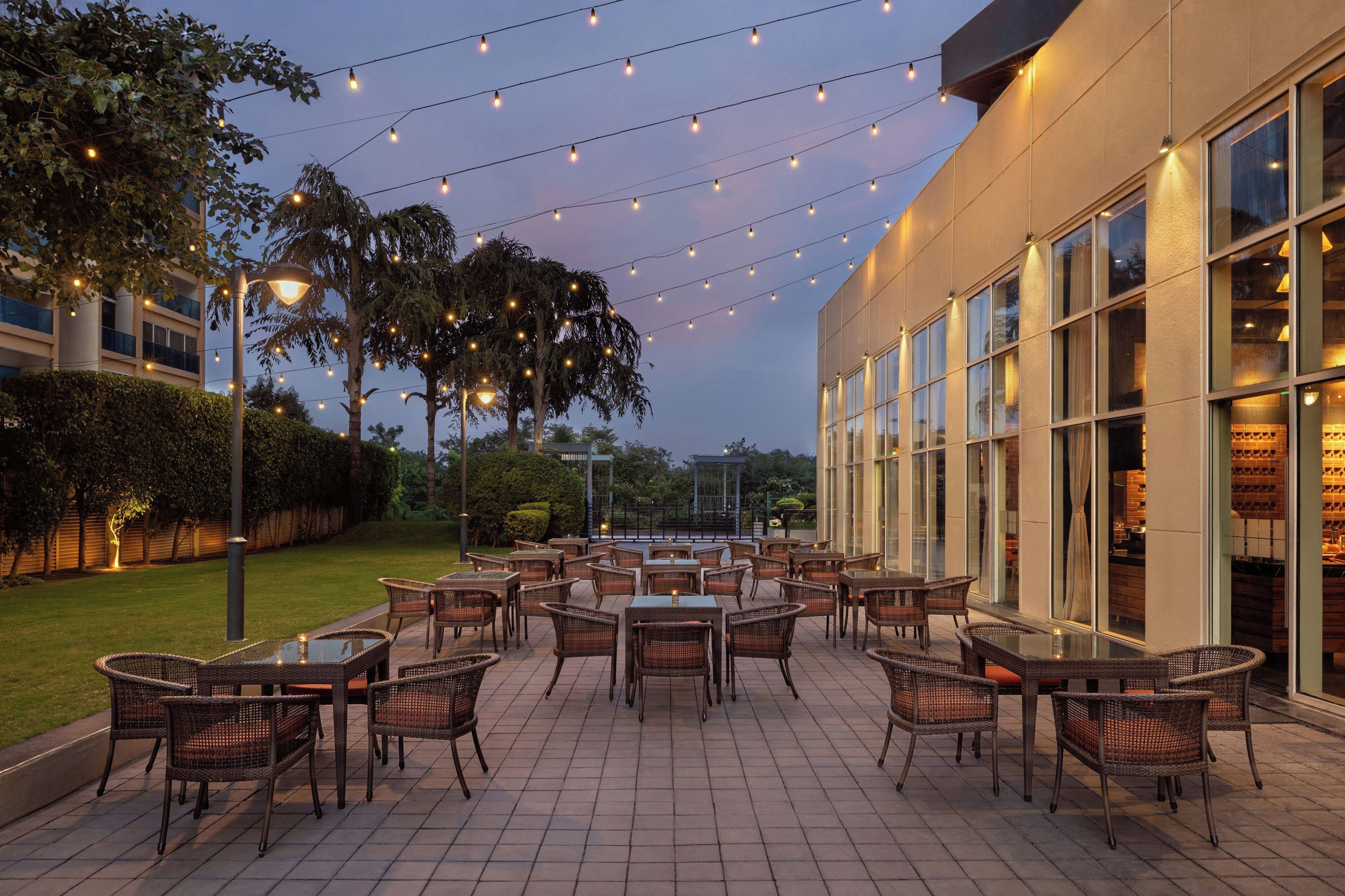 Un patio al aire libre con mesas y sillas, luces colgantes y un edificio con grandes ventanales.