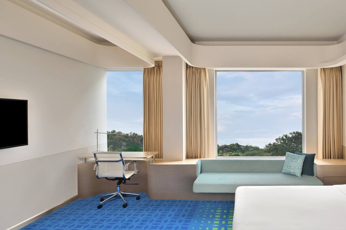 Ein Hotelzimmer mit Bett, Schreibtisch mit Stuhl, Fensterbank mit Kissen und einem großen Fenster mit Blick ins Freie.