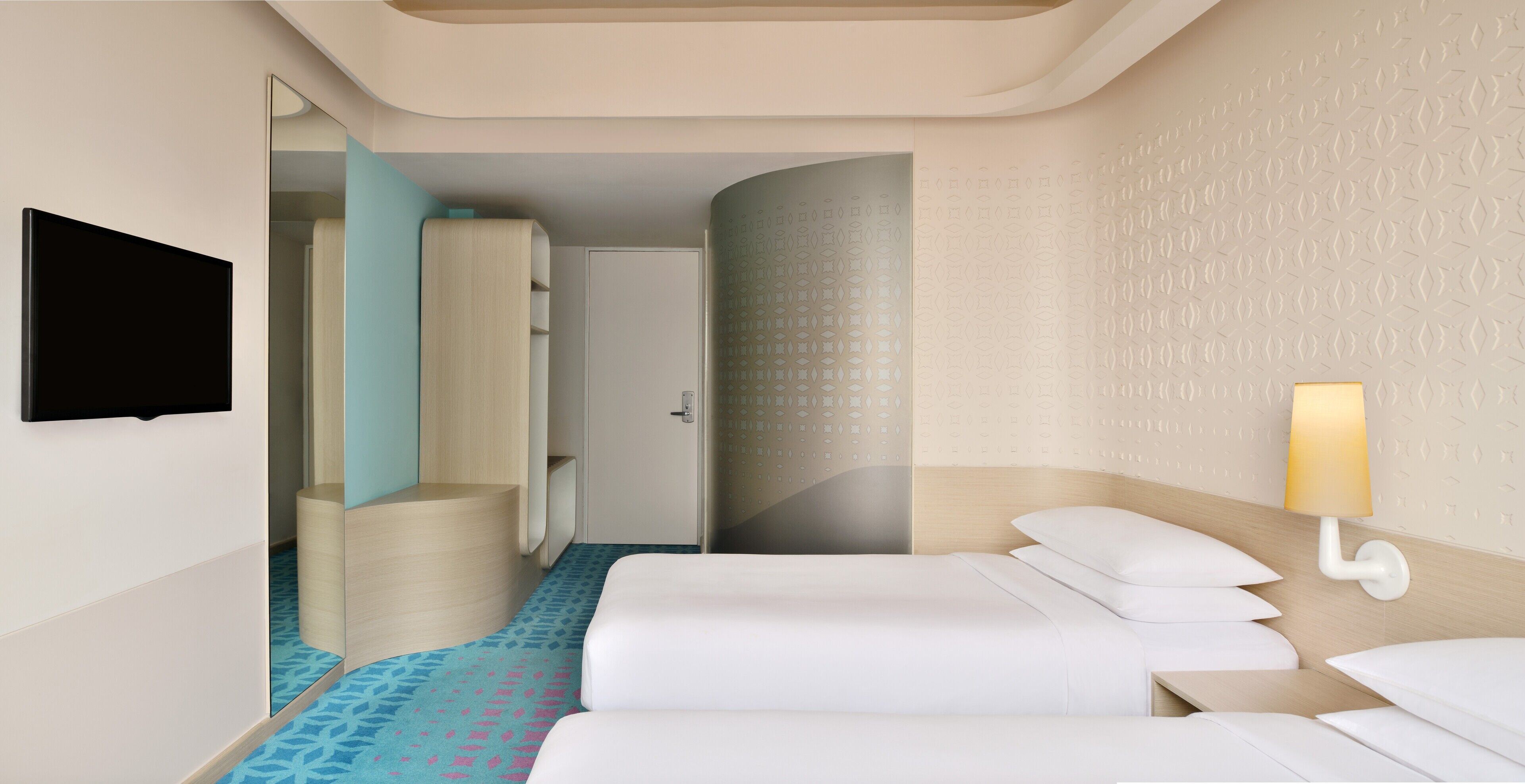 A hotel room with a bed, a TV mounted on the wall, a bedside lamp, and a patterned carpet.
