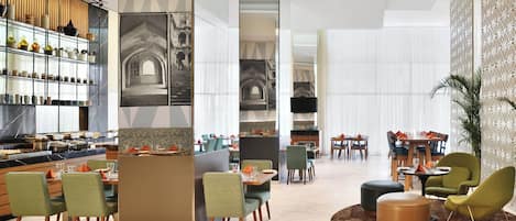 A modern hotel lobby with a dining area, a kitchen, and a seating area with chairs and a round table.
