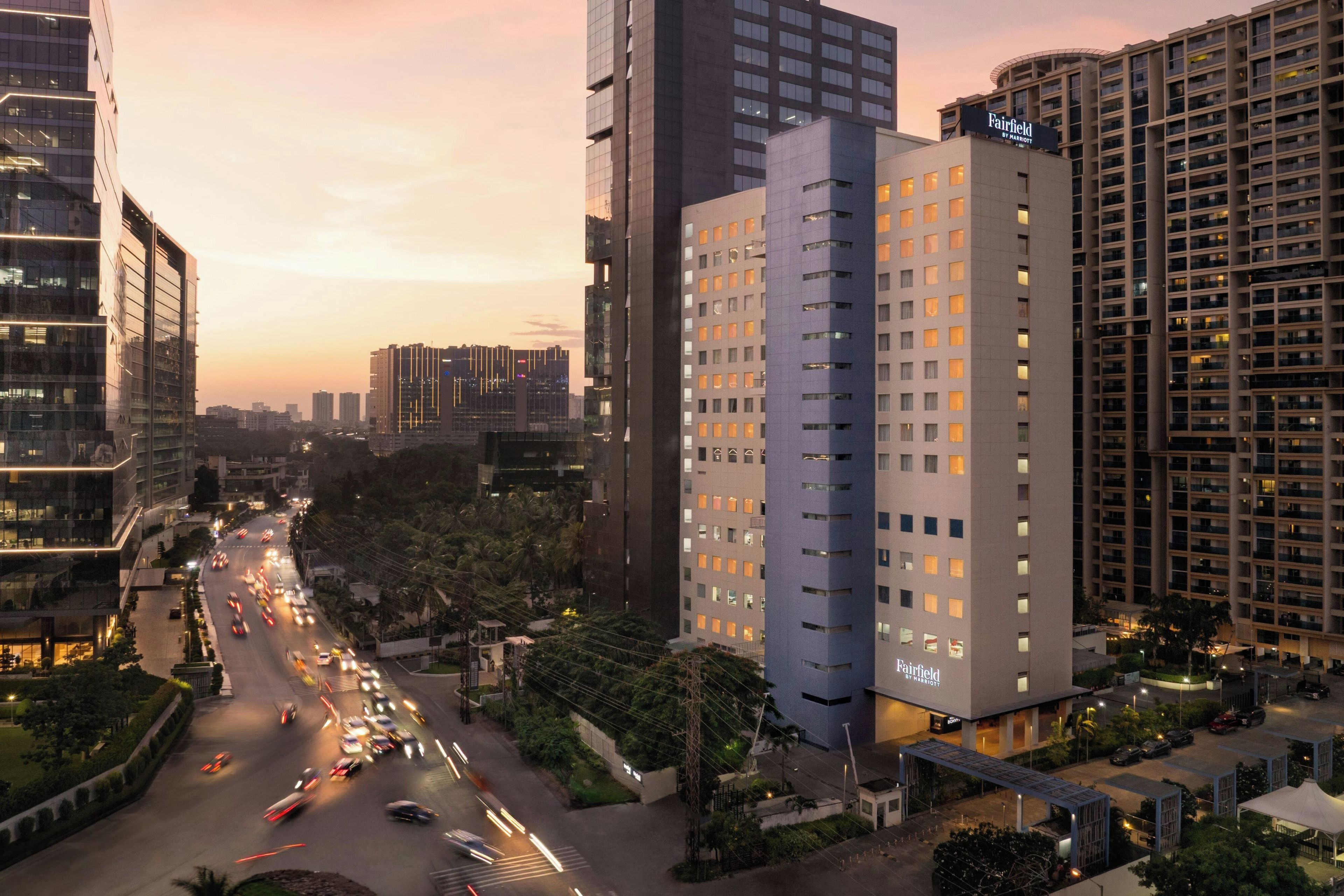 Un hotel de varios pisos con un llamativo letrero de 'Fairfield by Marriott', rodeado de edificios altos y una calle concurrida con vehículos en movimiento.