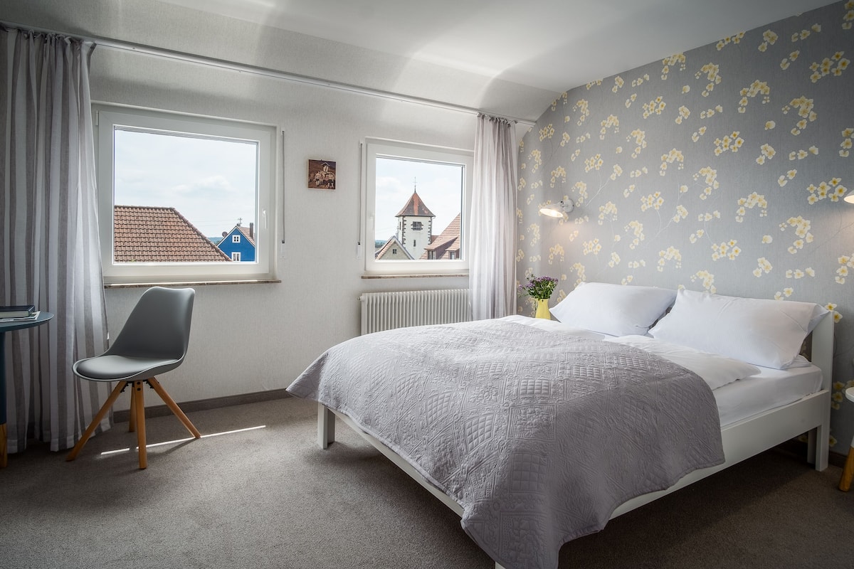 A bedroom with a bed, a chair, a nightstand, and two windows offering views of buildings.