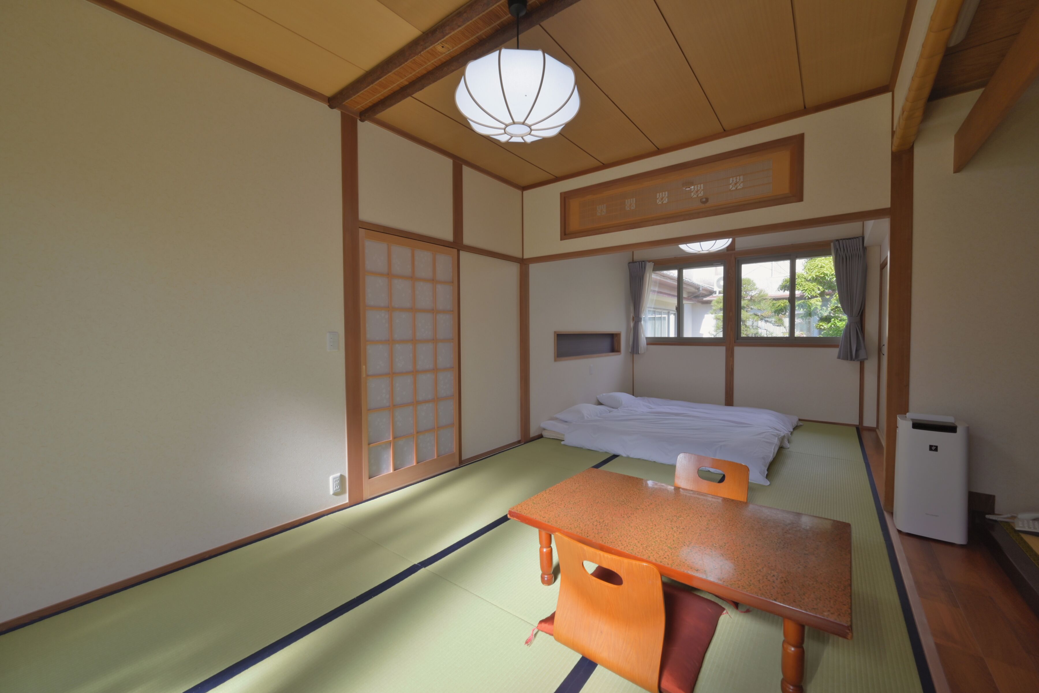 A traditional Japanese room with tatami flooring, a low wooden table, a bed with white bedding, and a ceiling light fixture.