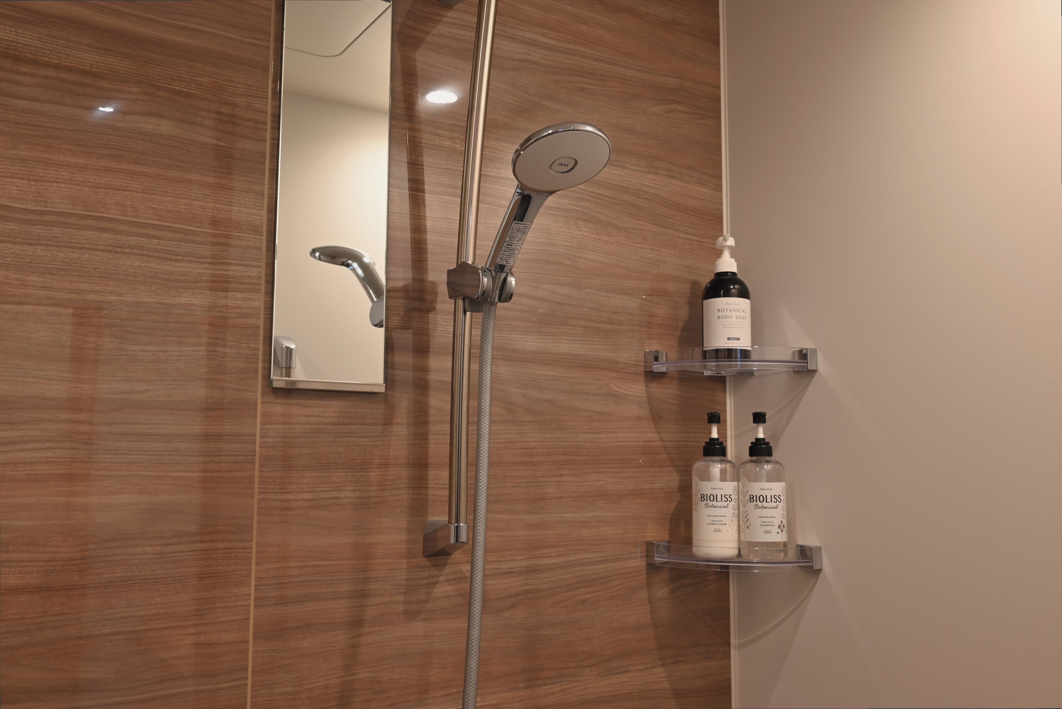 A modern bathroom with a wooden wall, a handheld showerhead, and two Bioliss body wash bottles on a shelf.