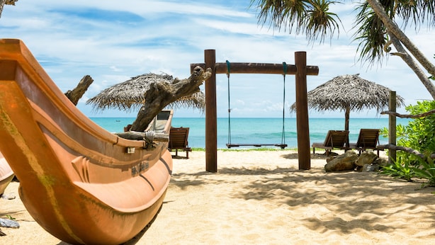 Une plage avec une balançoire en bois, un parasol en chaume et un grand pot en terre cuite.