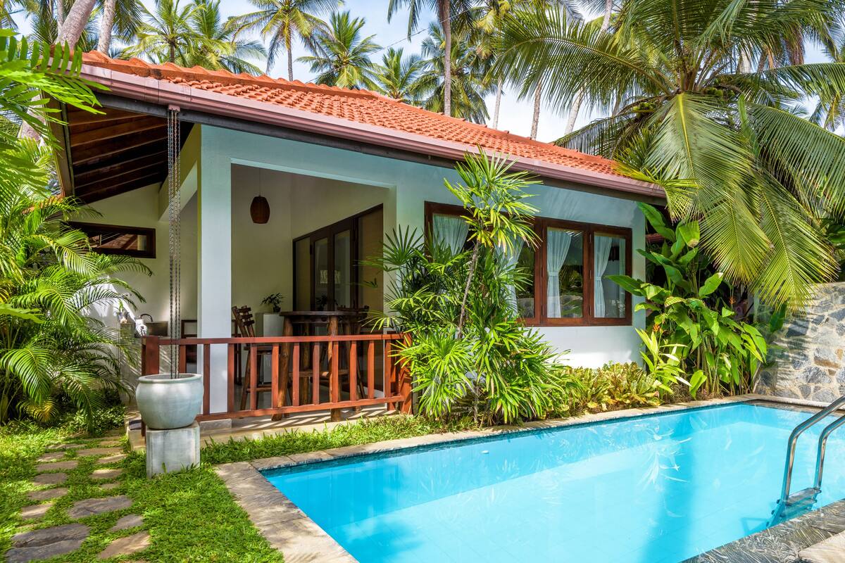 A two-story house with a swimming pool, surrounded by lush greenery and palm trees.