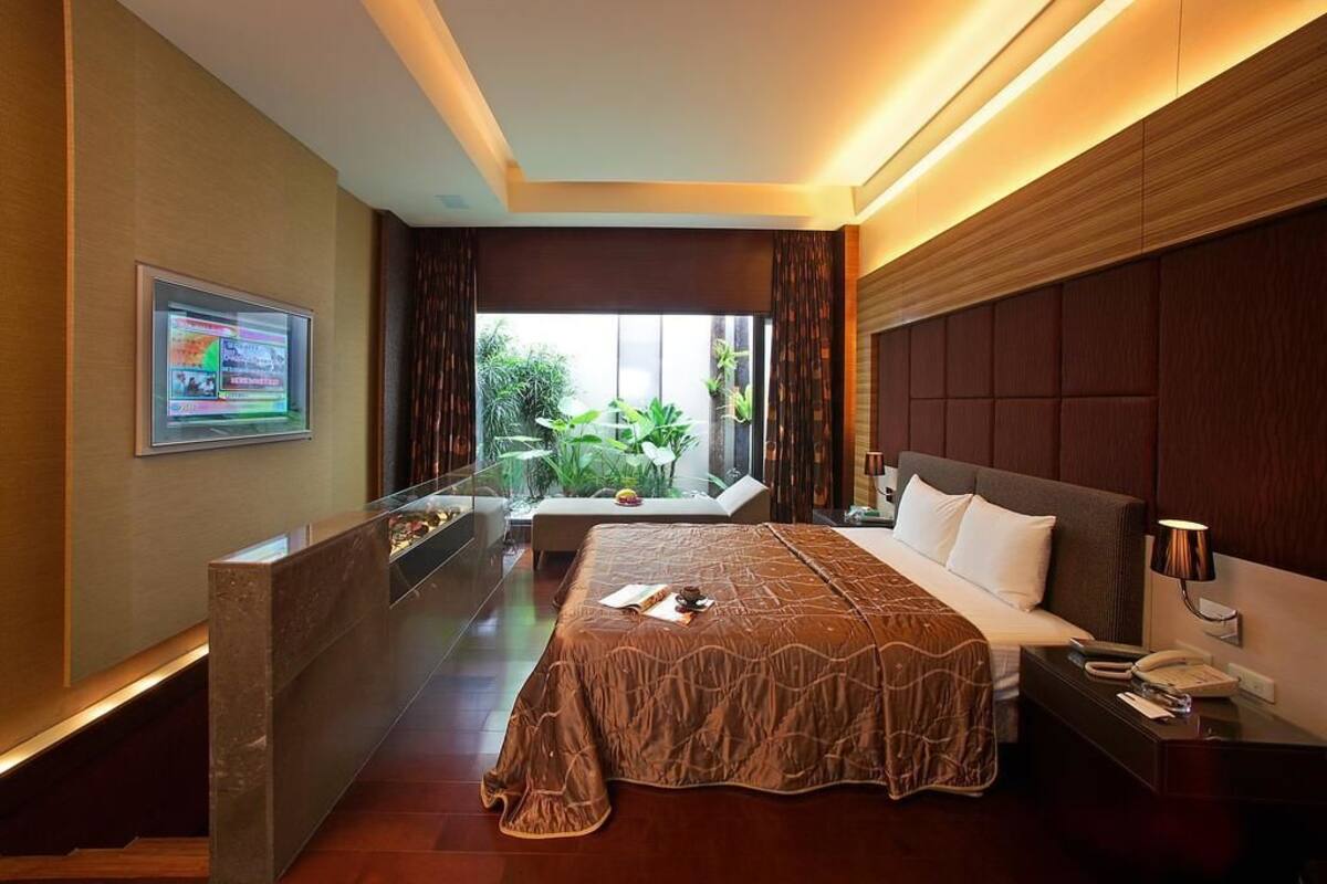 A hotel room with a large bed, a bedside table with a lamp, a TV mounted on the wall, and a view of greenery outside.