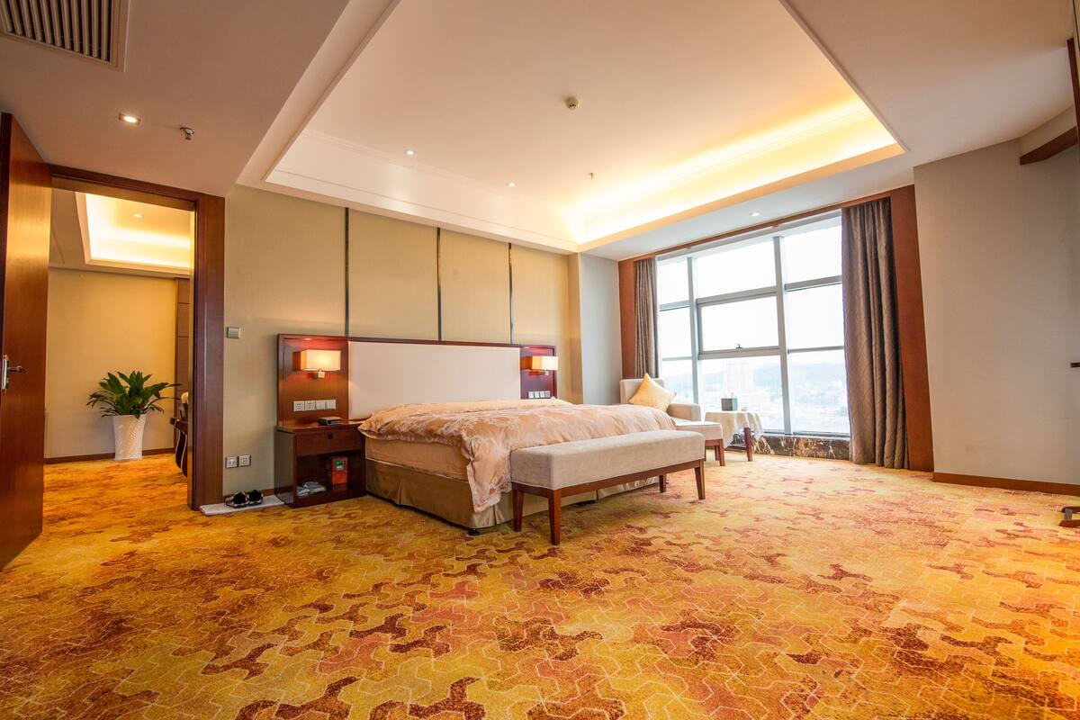 A hotel room with a large bed, a bench, a nightstand, a potted plant, and a large window with curtains.