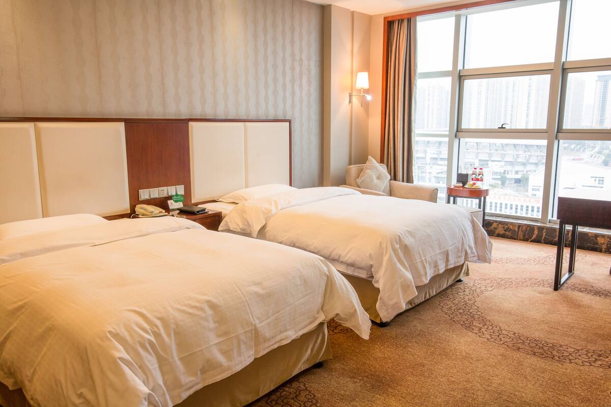 A hotel room with two beds, a nightstand, a lamp, a chair, and a view of the city through large windows.