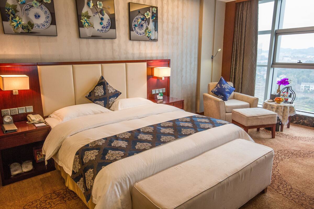 A hotel room with a large bed, a chair, a small table, and a view of the city through large windows.