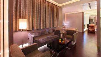 A modern hotel room with a sofa, armchairs, a glass coffee table, and a large window with curtains.