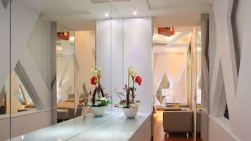A modern hotel corridor with a reflective floor, a potted plant with flowers, a seating area, and a digital control panel on the wall.