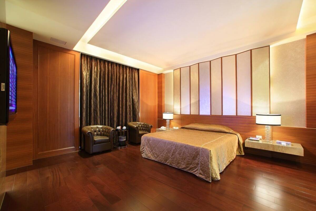 Quarto de hotel com uma cama grande, duas poltronas, uma televisão e piso de madeira.