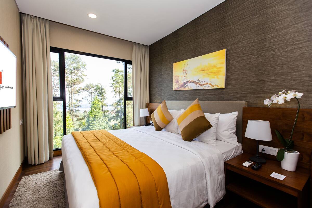Habitación de hotel con una cama grande, una mesita de noche con lámpara, una planta en maceta y un cuadro en la pared.