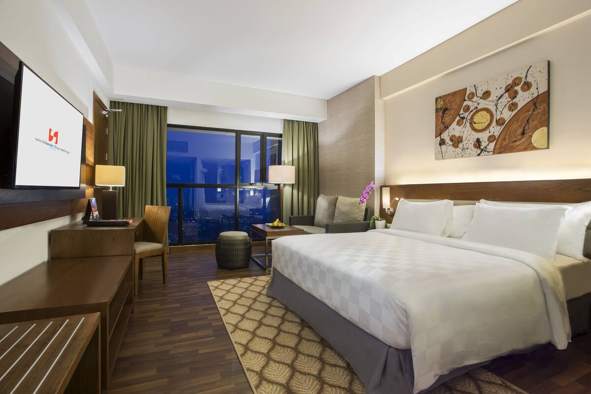 Habitación de hotel con una cama grande, un escritorio, una silla, una mesita y un ventanal con vistas a la ciudad.