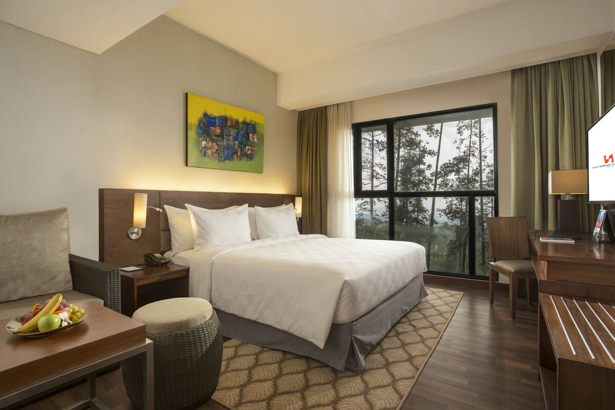 Habitación de hotel con una cama grande, un escritorio, una silla, una mesita con fruta, una lámpara y un cuadro en la pared.