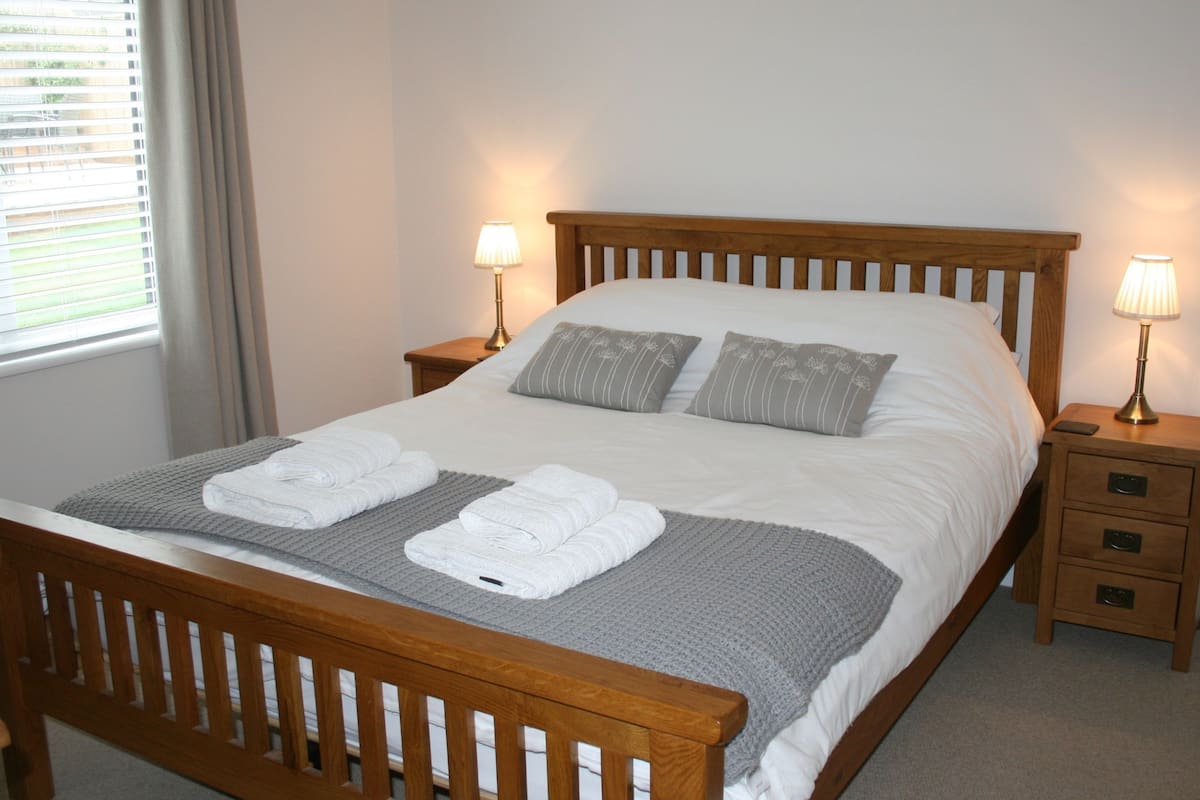 Una cama bien hecha con sábanas blancas, dos almohadas y una manta gris. Hay dos mesitas de noche con lámparas, y un tocador de madera con una lámpara.