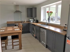 Une cuisine moderne avec des meubles gris, une table à manger en bois, un lave-linge encastré et une fenêtre agrémentée d’un vase de fleurs.