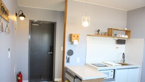 Uma cozinha compacta com geladeira, micro-ondas e pia. Há uma prateleira de madeira acima da pia e um extintor próximo à porta.