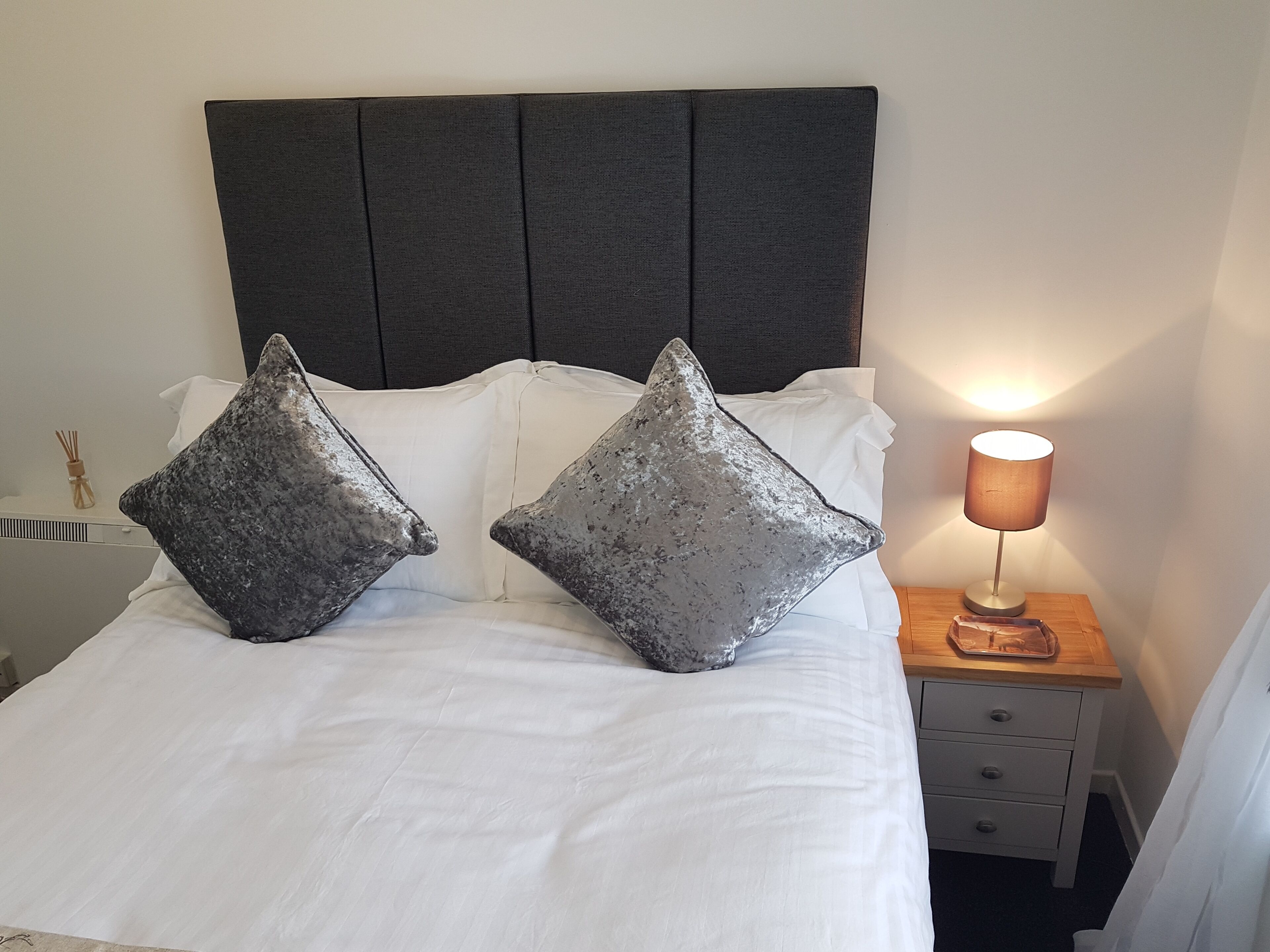 A bed with white bedding and two metallic pillows, a bedside lamp, and a wooden nightstand with drawers.