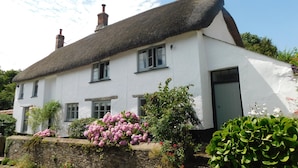A white building with a thatched roof, multiple windows, and a stone wall with flowering plants. - Middletown Farmhouse B&B (Okehampton)