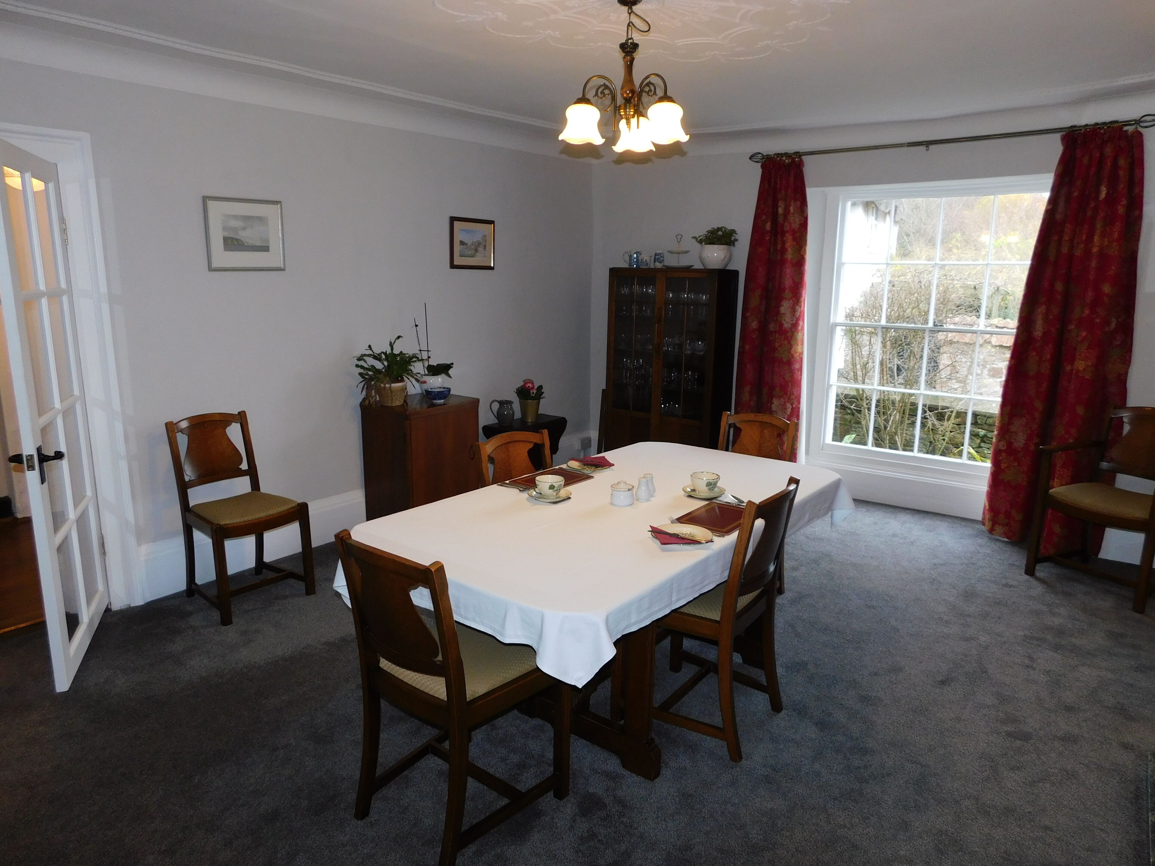 A dining room with a table set for four, surrounded by wooden chairs. There is a chandelier above the table, a window with curtains, and a cabinet with glass doors.