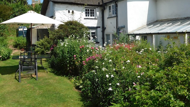 A traditional thatched-roof house with a white exterior, surrounded by lush greenery and flowering plants. - Middletown Farmhouse B&B (Okehampton)