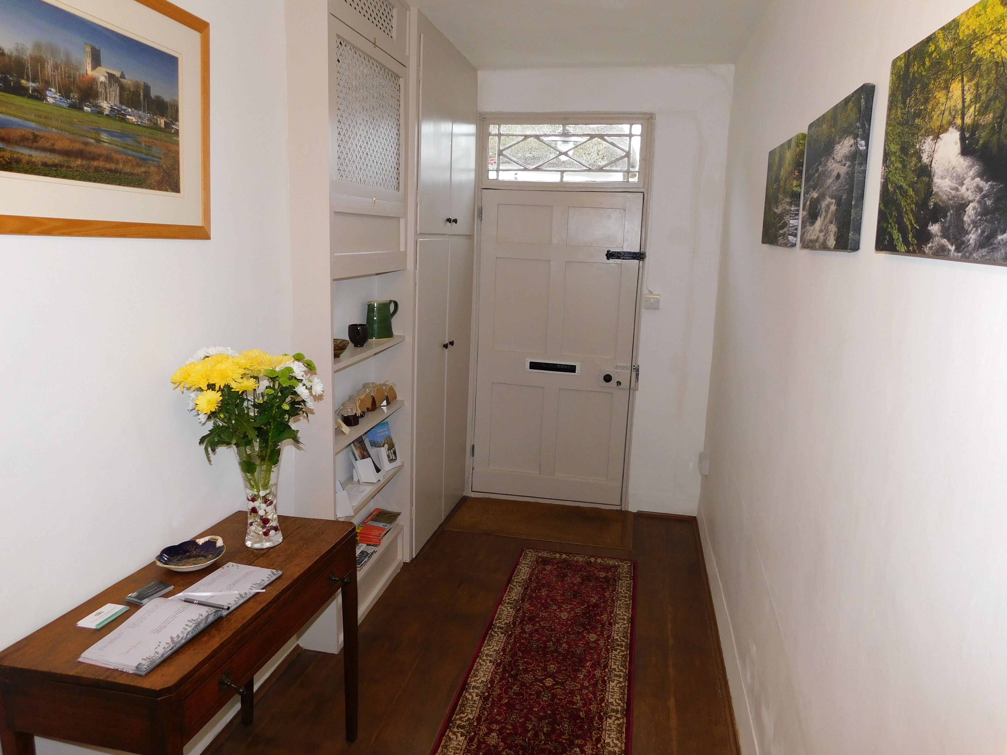A hallway with a wooden desk, a vase of flowers, a bookshelf, and a door leading to another room.