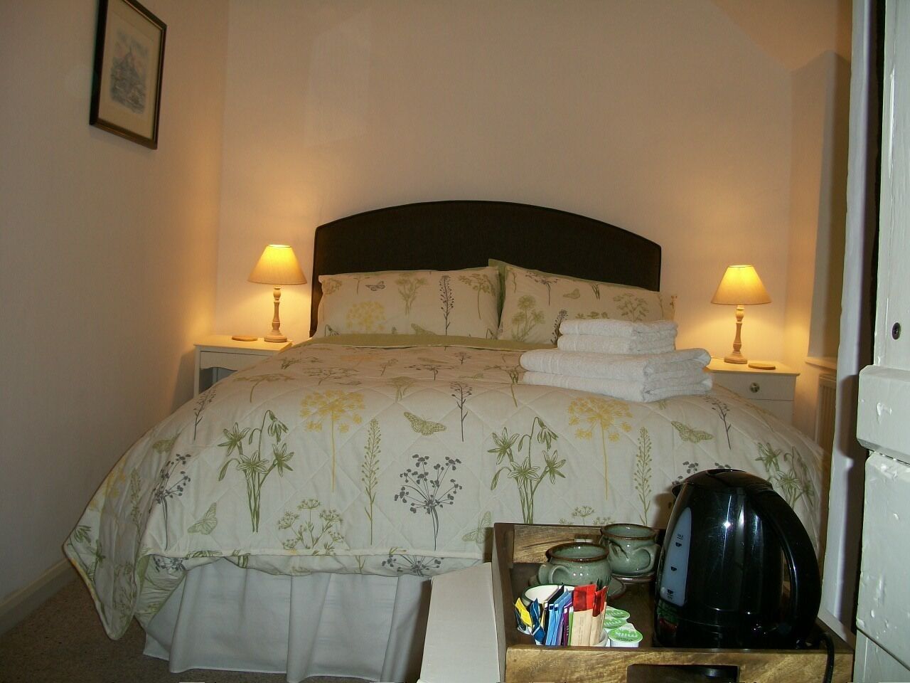 A neatly made bed with floral bedding, two bedside lamps, a small table with a kettle and books, and a framed picture on the wall.