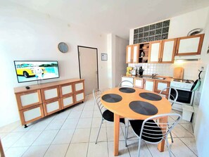 A compact kitchen with a dining area, a clock, and a television displaying a car scene.