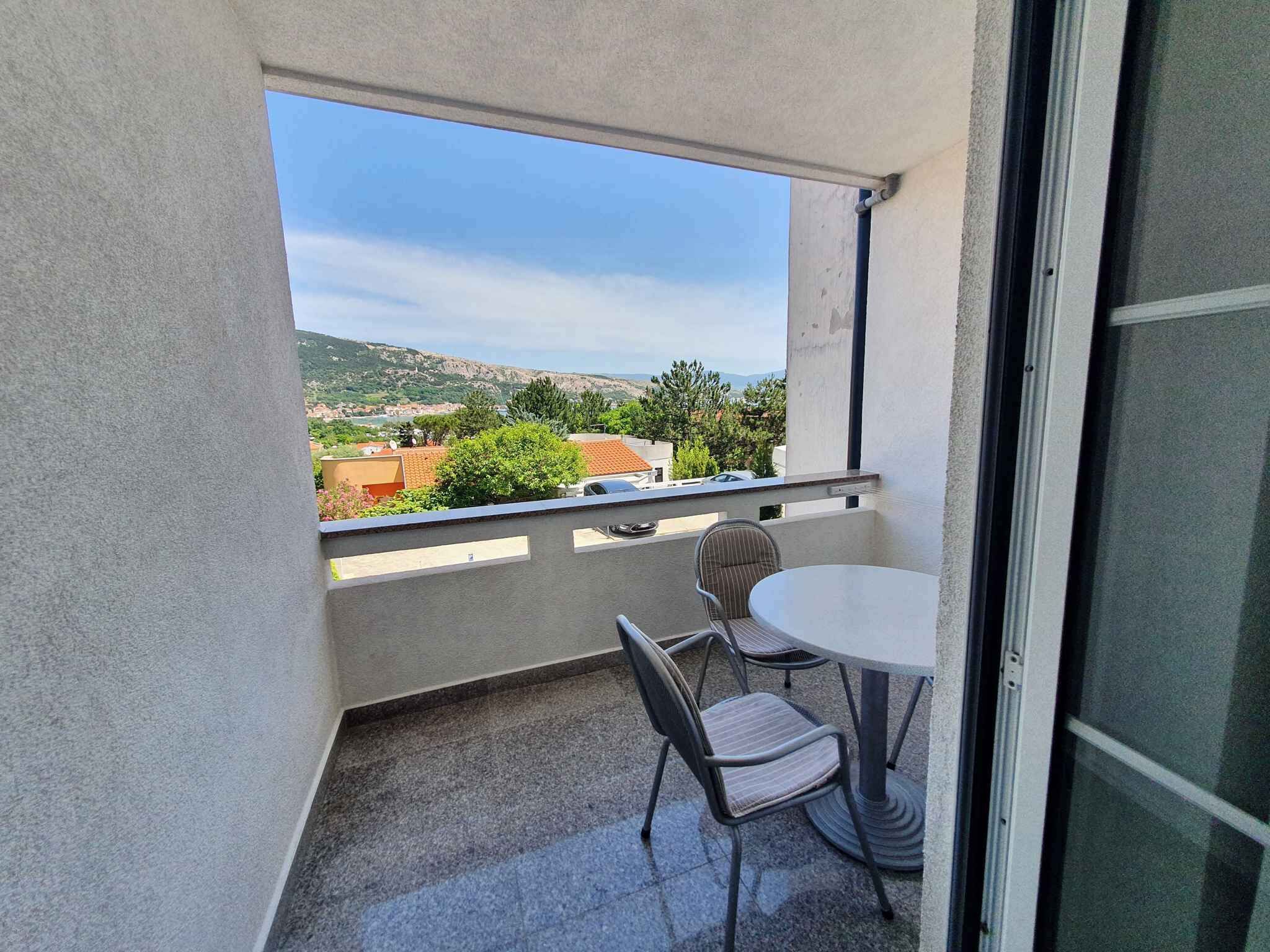 A balcony with a table and two chairs, a view of a hilly landscape, and a clear sky.