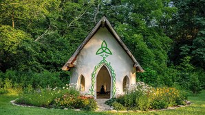 Una pequeña estructura de techo triangular blanco con un símbolo de la Santísima Trinidad verde, rodeada de un jardín con diversas plantas y flores.