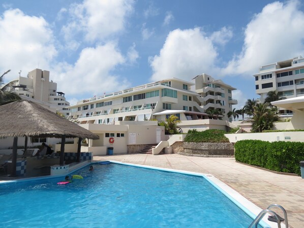 A swimming pool with a thatched roof structure, surrounded by multi-story buildings with balconies and a clear sky. - Wake up in Caribbean Paradise 1304 (Cancún)