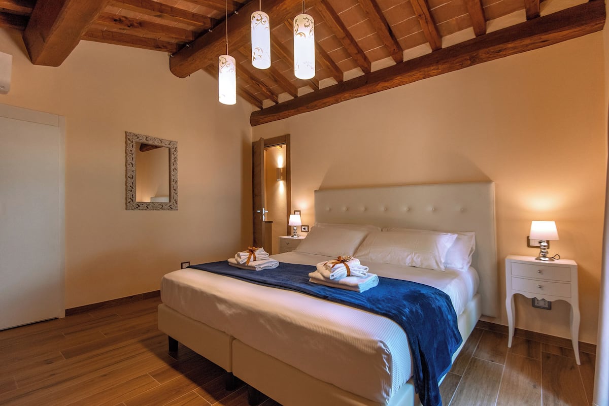 Een slaapkamer met een groot bed, houten balken in het plafond, een spiegel, bedlampen en een keurig opgemaakt bed met handdoeken.
