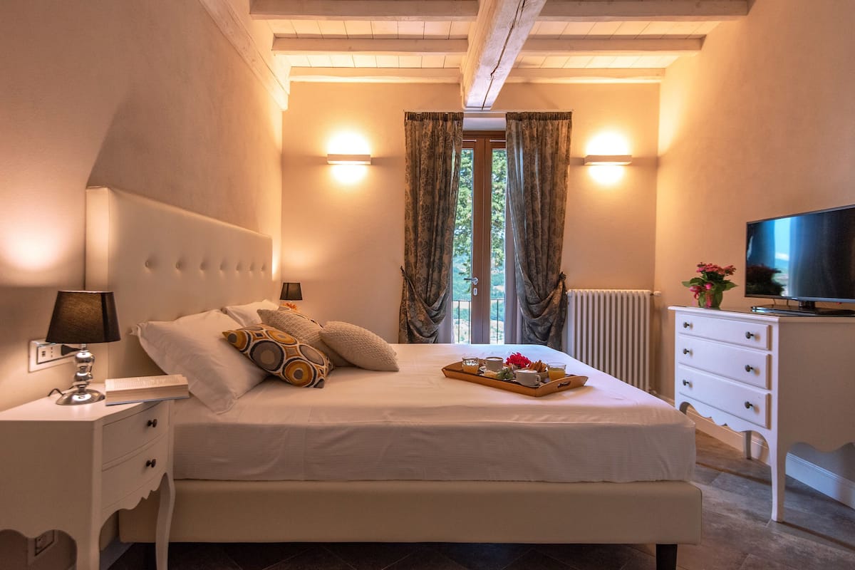Een slaapkamer met een bed, nachtkastje, lamp, dienblad met ontbijtartikelen, gordijnen, raam en een televisie op een dressoir.