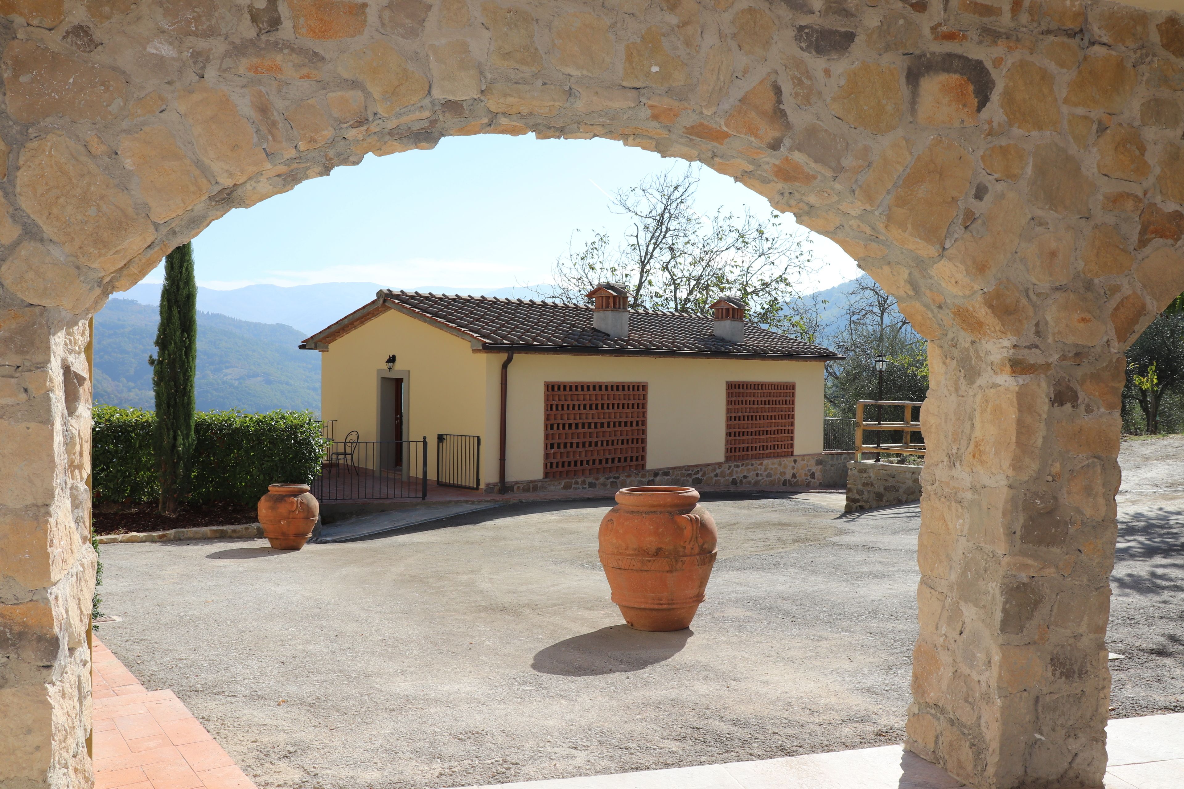 A stone archway leading to a small building with a tiled roof, surrounded by a paved area and two large terracotta pots.
