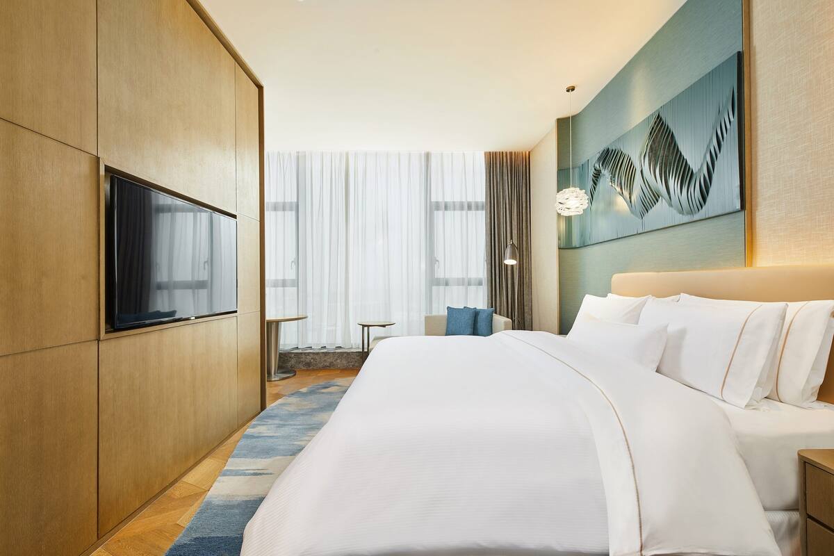 A modern hotel room with a large bed, a flat-screen TV, a decorative wall piece, and a view of the outdoors through large windows.