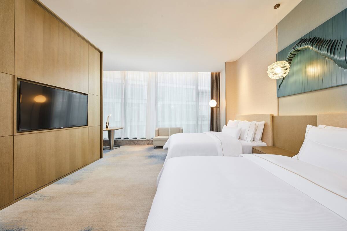 A hotel room with two beds, a TV, a desk, and a large window.