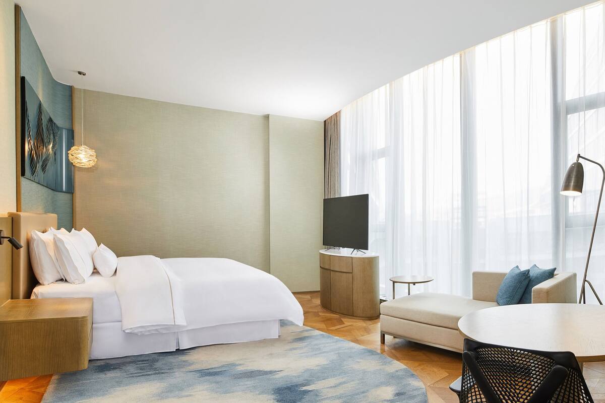 A modern hotel room with a bed, a TV, a sofa, a small table, and a chair.