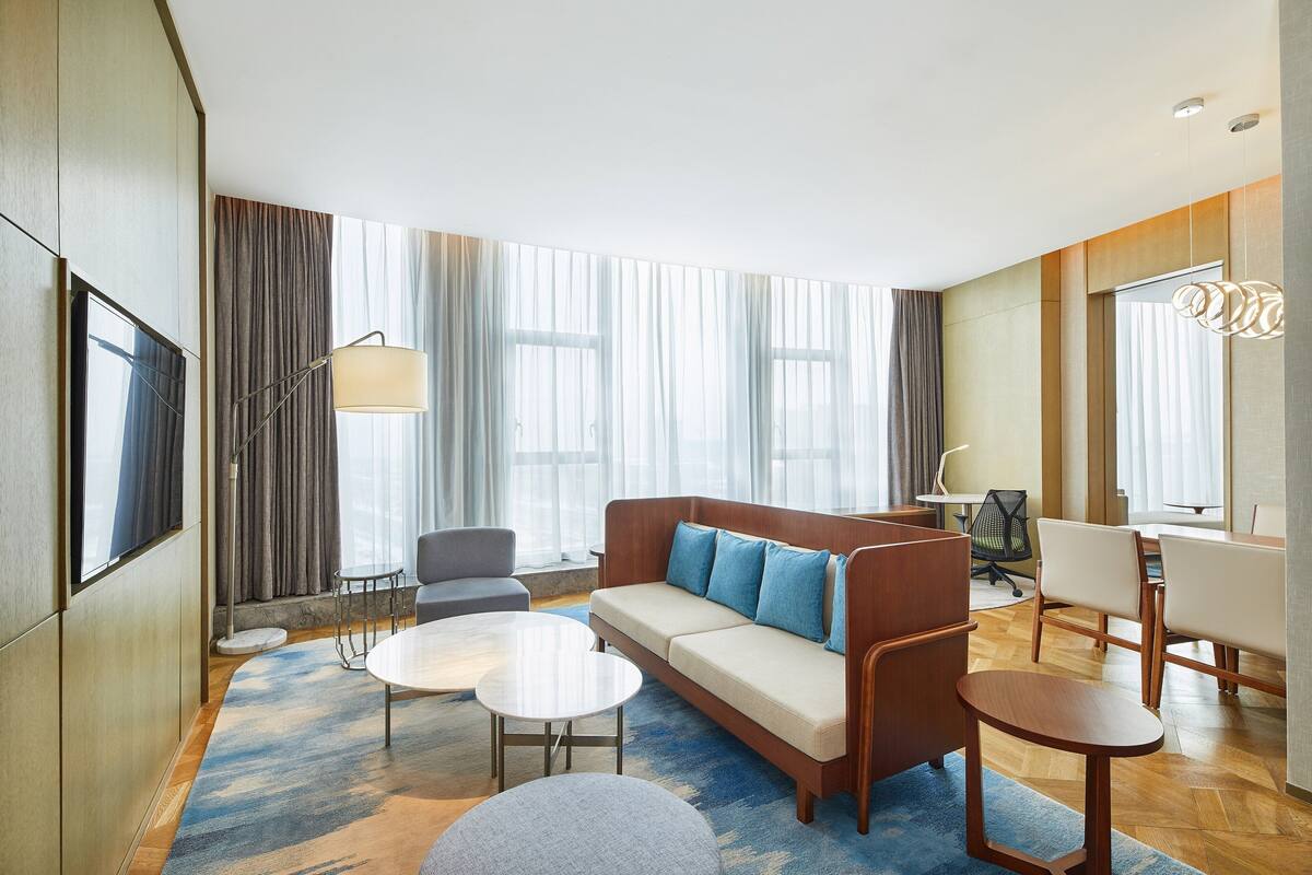 A modern hotel room with a sofa, two armchairs, a coffee table, a desk, and a TV.
