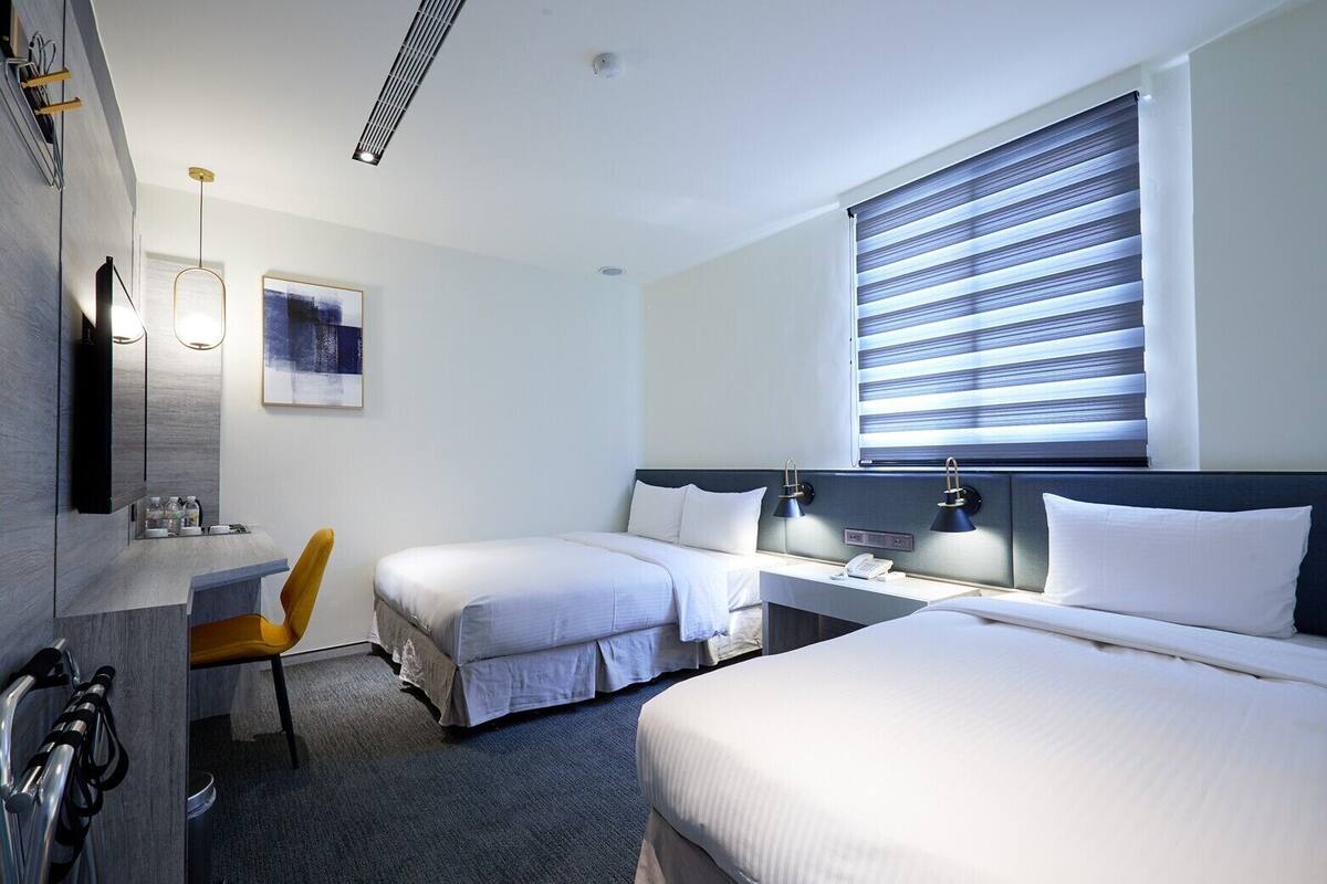 Une chambre d’hôtel avec deux lits, un bureau, une chaise, un tableau et une fenêtre avec des stores.