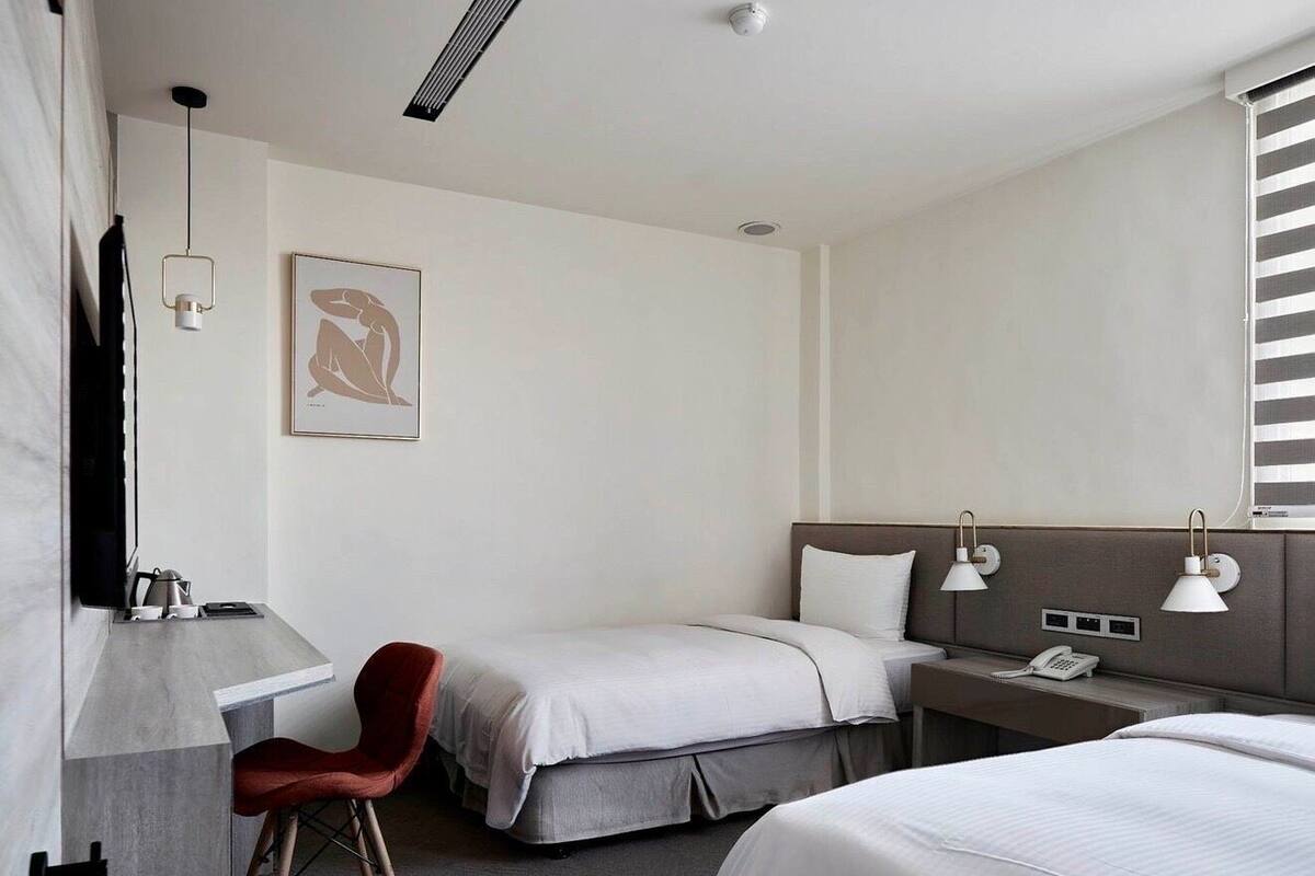 Une chambre d’hôtel avec deux lits, un bureau, une chaise, une table de chevet avec une lampe, un téléphone et un tableau au mur.