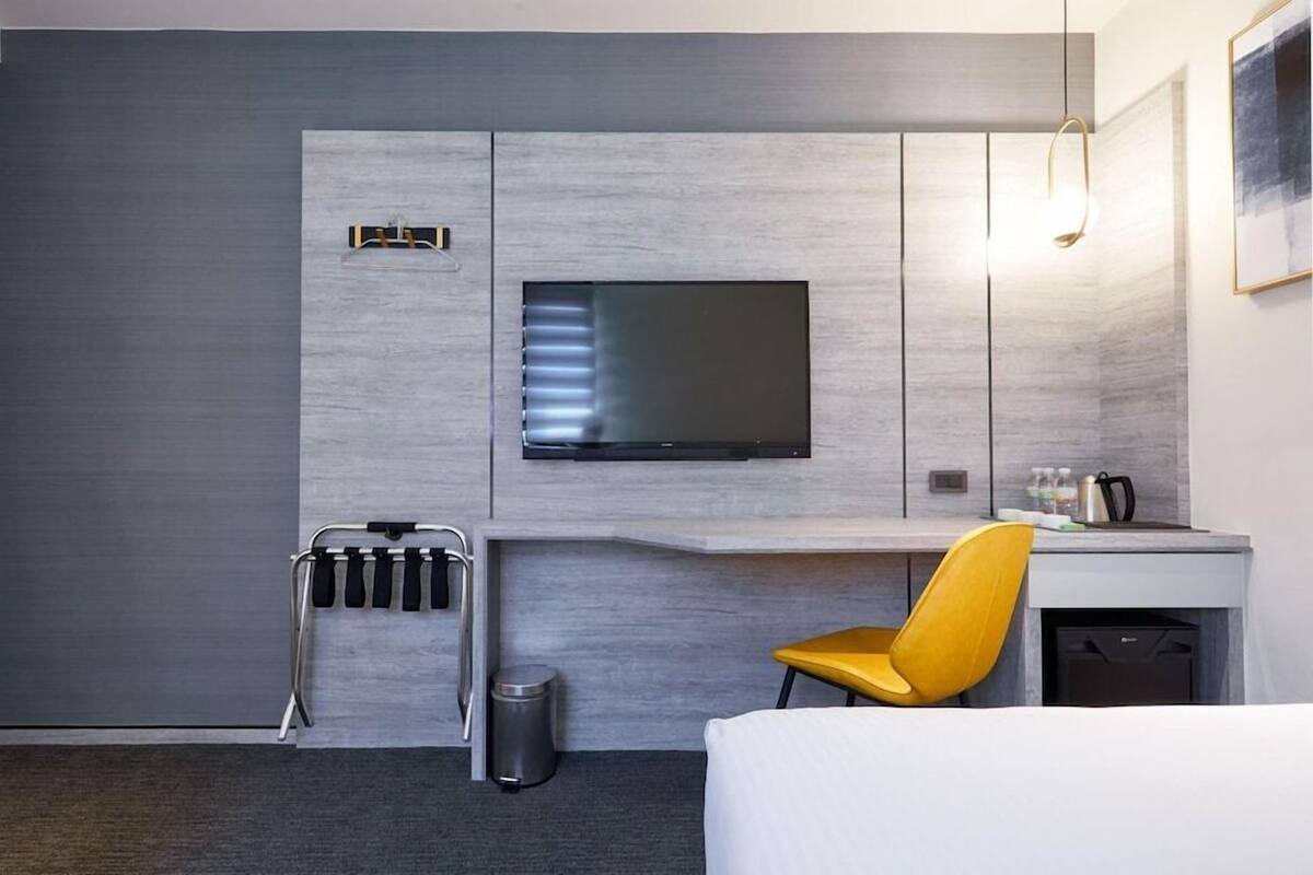 Une chambre d’hôtel moderne équipée d’un téléviseur à écran plat fixé au mur, d’un bureau avec une chaise, d’une poubelle et d’un lit.