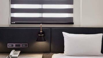 Habitación de hotel con cama, mesita de noche con teléfono, lámpara y ventana con persianas horizontales.