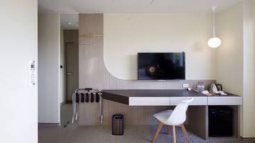 Una habitación de hotel moderna con un televisor de pantalla plana montado en la pared, un escritorio pequeño con una silla, un perchero y un cesto de basura.