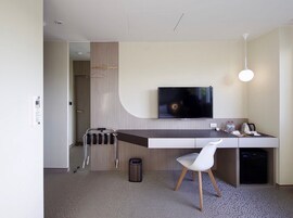 Una habitación de hotel moderna con un televisor de pantalla plana montado en la pared, un escritorio pequeño con una silla, un perchero y un cesto de basura.