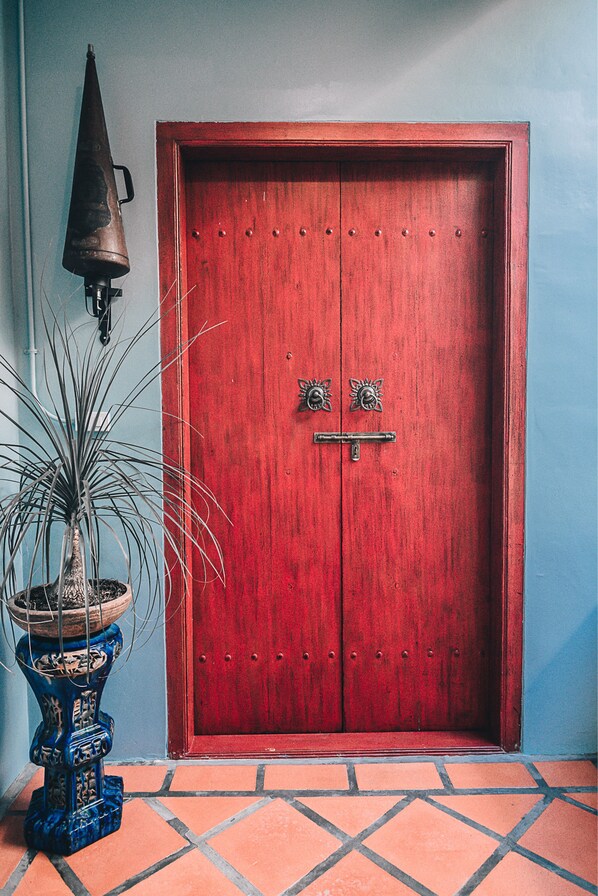 A red wooden door with two handles and a metal knocker, a blue ceramic pot with a plant, and a tiled floor. - Woo Gallery and Boutique Hotel (Phuket)