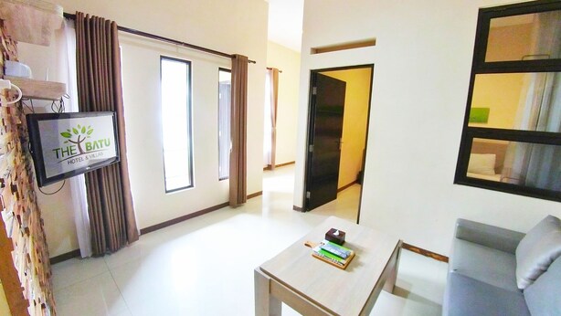 A hotel room with a flat-screen TV displaying 'The Batu Hotel & Villas' sign, a sofa, a coffee table, and a door leading to another room. - Villa Batu 2 kamar 5 dekat Museum Angkut (Batu)